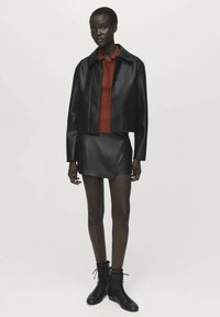 Zwarte nep leren cropped jack, rode geribde top, zwarte mini rok en zwarte veterschoenen. Model staat tegen een effen achtergrond.