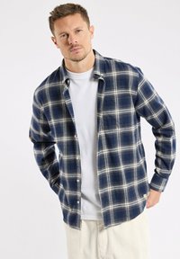 Homme portant une chemise à carreaux bleu marine et blanche par-dessus un t-shirt blanc et un pantalon beige, se tenant debout devant un fond clair.