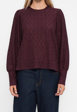 Kobieta w bluzce z ciemnofioletowej koronki z długim rękawem i bufkami na ramionach, w połączeniu z ciemnoniebieskimi jeansami z wysokim stanem.