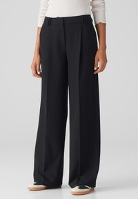Black wide-leg trousers with a tailored design, pleated front, and a high waistband. Smooth fabric with a crisp finish. Shoes: white and black.