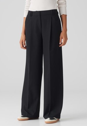 Schwarze weite Hosen mit tailliertem Design, gefalteter Vorderseite und hohem Bund. Glattes Material mit einem klaren Finish. Schuhe: schwarz und weiß.