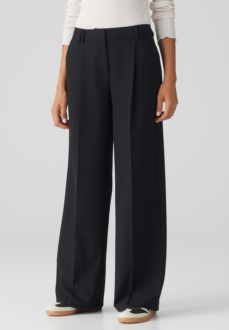 Black wide-leg trousers with a tailored design, pleated front, and a high waistband. Smooth fabric with a crisp finish. Shoes: white and black.