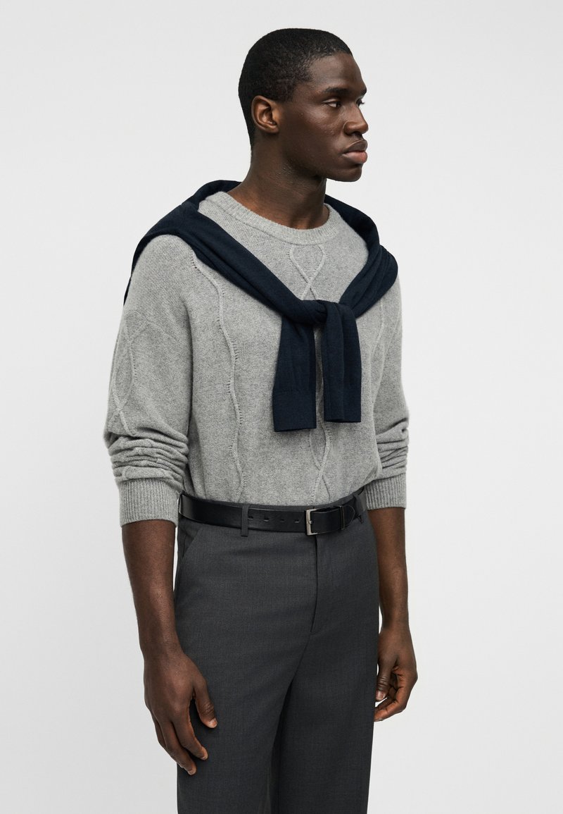 Pull gris à maille torsadée avec une écharpe bleu marine drapée sur les épaules, associé à un pantalon sur mesure gris foncé et une ceinture noire.