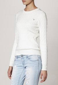 Witte kabelbreipullover met een ronde halslijn en geribbelde boorden, gecombineerd met lichtblauwe distressed jeans. Heeft een logo op de borst.