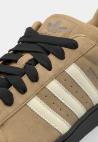 Brown suede sneaker with black laces, cream stripes, perforated detailing, and a gray logo patch on the tongue. Black rubber outsole.