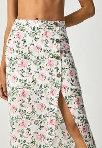 Jupe midi florale en tissu blanc avec des motifs de fleurs roses et vertes. Elle présente une fente sur le côté et cinq boutons décoratifs à l'avant.