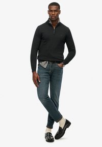 Zwarte zip-hals trui, geruite shirt eronder, blauwe spijkerbroek, lichtgekleurde sokken, zwarte instappers. Model poseert met gekruiste armen.