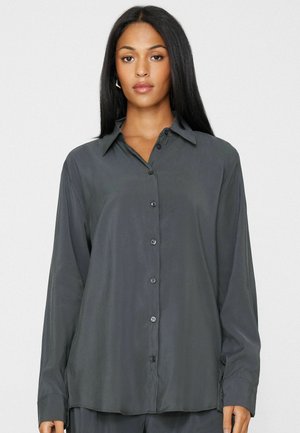Donna con lunghi capelli neri che indossa una camicia grigio scuro con bottoni e colletto, in piedi davanti a uno sfondo chiaro e semplice.