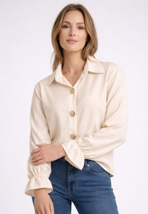 Frau trägt cremefarbene Bluse mit goldenen Knöpfen und Rüschen an den Manschetten, kombiniert mit blauen Jeans, posiert vor weißem Hintergrund.