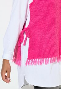 Heldere roze gebreide vest met franjes, voorzien van zijstrikken. Draag het over een wit hemd, waarop een textuurpatroon zichtbaar is.
