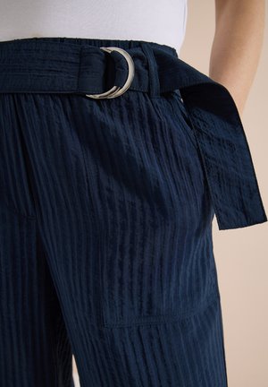 Personne portant un pantalon bleu marine texturé avec une grande poche plaquée et une ceinture assortie en tissu avec boucle anneau en D argentée.