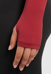 Rode fitted mouw met duimopening, gemaakt van glad, stretchstof. Kenmerkt zich door nette stiksels en een nauwsluitende pasvorm tegen de huid. Achtergrond is zwart.