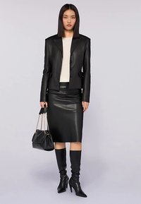 Schwarze Lederjacke mit scharfem Kragen, kombiniert mit einem taillierten schwarzen Lederrock. Das Modell hält eine schwarze Tasche und trägt kniehohe schwarze Stiefel.