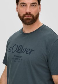 Vyrų apvalaus kaklo t-shirt tamsiai žalia spalva, pagamintas iš medvilnės. Su dideliu tamsiai mėlynos spalvos grafiniu įvaizdžiu, kuriame yra prekės ženklas ir šūkis, centruotas krūtinėje. Trumpomis rankovėmis.