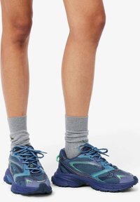 Scarpe sportive in tonalità di blu con pannelli in mesh, suole testurizzate e dettagli verdi. Abbinate a calzini alla caviglia grigi a costine.