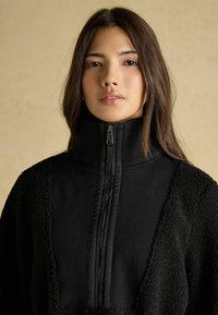 Młoda kobieta z prostymi brązowymi włosami, ubrana w czarną kurtkę o teksturowanej powierzchni z wysokim suwakowym kołnierzem, patrzy bezpośrednio przed siebie.