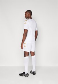 Hvid fodboldtrøje og shorts med gyldne Kappa-logo accenter på skuldrene. Sorte fodboldsko og hvide sokker. Minimalistisk design.