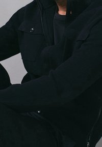 Man i svart jacka och svart skjorta, sittande med armarna i kors, mot en enfärgad grå bakgrund.