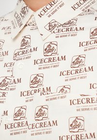 Hvit skjorte med gjentatt brun "ICECREAM" logo og tegneserie-is karakter. Har spiss krage og knappeåpning.