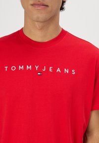 T-shirt en coton rouge avec un col rond, arborant le logo "TOMMYJEANS" en blanc, accompagné d'un petit emblème de drapeau bleu et rouge.