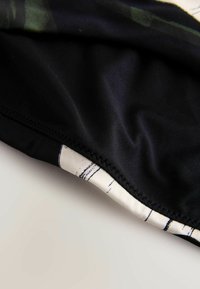 Melnā auduma virsma ar gludu tekstūru, kas rotāta ar krēmkrāsas rakstu malu ar melnām līnijām. Dizainā ir redzamas smalkas krāsu variācijas.