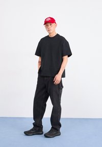 Ung man med tatueringar, iklädd en röd keps, svart oversized t-shirt, lösa svarta jeans och svarta sneakers, står på ett blått golv mot en vit vägg.