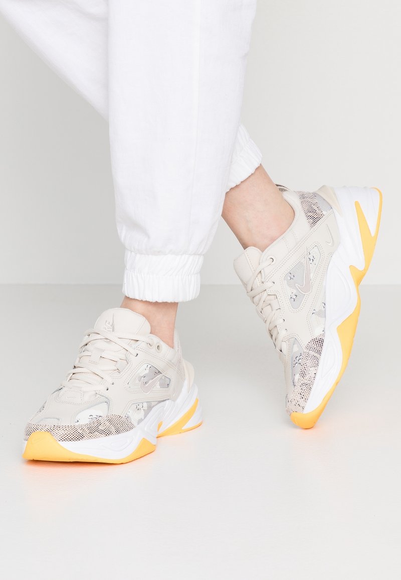 Personne portant des baskets blanches et beiges avec des semelles orange, associées à un pantalon de jogging blanc sur un fond clair uni.