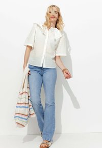Blouse blanche en dentelle avec manches courtes, col froncé et fermeture à boutons ; associée à un jean flare bleu clair et un sac en crochet multicolore.