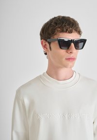 Occhiali da sole neri e angolari con lenti scure, indossati con una felpa crema caratterizzata da un logo embossato. Texture liscia ed elementi di design minimi.