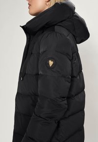 Kvinna som bär en svart kviltad pufferjacka med hög krage och huva, med ett cirkulärt märke med ett guldhjärta på ärmen.