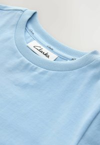 Ljusblå bomullströja med ribbad rundad halsringning, med en Clarks-märke sydd på baksidan. Slät yta och enkel design.