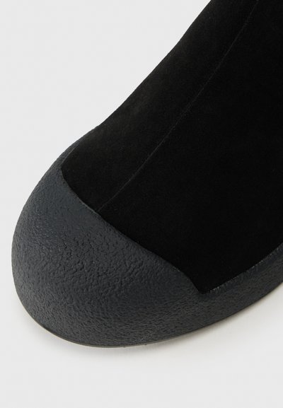 Juodas suède kulkšninis batas su tekstūriniu guminio bato galvute. Dizainas apima lygią šoninės dalies panelę ir apvalią bato formą.