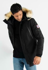 Parka noire avec capuche en fourrure, plusieurs poches zippées, tissu texturé et cordons de serrage réglables. Logo brodé sur la manche.