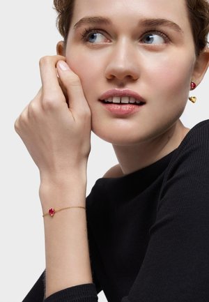 Pulsera de oro con una pequeña gema roja en forma de corazón, montada en una delicada cadena, llevada en una muñeca. El modelo muestra un primer plano de la joyería.