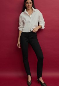 Camisa blanca recortada de botones con mangas enrolladas, combinada con pantalones negros ajustados y tacones negros transparentes, contra un fondo rojo.