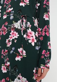 Bloemrok met een donkergroene achtergrond, pink en witte bloesems, lange mouwen en een tailleband. Zachte, lichte stof.