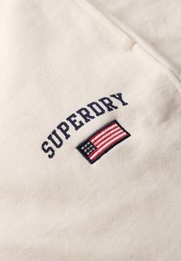 Crème-kleurige sweatshirt met "SUPERDRY" geborduurd in marineblauw bovenop een klein American flag patch in rood, wit en blauw. Zachte stoftextuur.