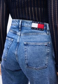 Άτομο που φοράει ψηλόμεσα ανοιχτό μπλε τζιν με λογότυπο Tommy Jeans στη μέση και μικρή σημαία στην πίσω τσέπη, συνδυασμένα με σκούρο υφασμάτινο τοπ.