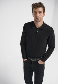 Svart långärmad pikétröja med krage och en treknappslinning. Bärs med mörka jeans och svart bälte, enfärgad bakgrund.