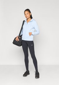 Ανοιχτό μπλε φούτερ με φερμουάρ, κουκούλα και δύο μπροστινές τσέπες, σε συνδυασμό με μαύρο κολάν με λογότυπο. Το μοντέλο κρατά μια μαύρη τσάντα και φοράει σκοτεινά παπούτσια.