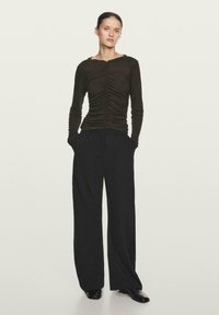 Maglia a maniche lunghe marrone con texture arricciata sul davanti, scollatura a barca, abbinata a pantaloni neri a gamba larga e scarpe nere.