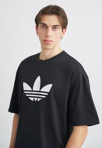 Black cotton t-shirt featuring a large white Adidas trefoil logo with three horizontal stripes beneath. Short sleeves and crew neckline.