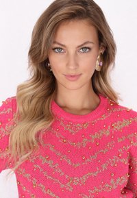 Světle růžový pletený svetr se zlatými korálkovými akcenty a zigzagovým vzorem. Modelka má na sobě velké zlaté a fialové náušnice. Hladká textura.