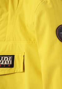 Gul jakke med et glat, letvægtsstof, en markant sort lommelap og en cirkulær logo-accent på ærmet.