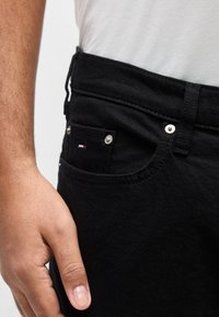 Κοντινό πλάνο ενός χεριού που ακουμπά κοντά στην εμπρόσθια τσέπη μαύρων τζιν με μικρό κόκκινο, άσπρο και μπλε λογότυπο και μεταλλικά καρφιά.