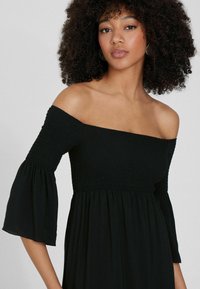 Vestido negro sin hombros con cuerpo fruncido y mangas acampanadas, que presenta una falda fluida y una textura suave. Sin patrones o acentos notables.