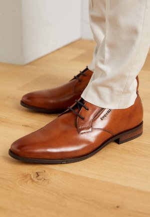 Scarpe in pelle marrone con una finitura lucida, design allacciato, punta arrotondata e dettagli di cucitura minimi. Indossate con pantaloni chiari.
