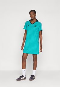 Tirkizna polo haljina s vertikalnim bijelim prugama, crnom ovratnikom i detaljima, te logotipom na prsima. Model nosi sive tenisice i bijele čarape.