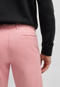 Pantalones de algodón rosa claro con corte recto. Cuentan con un bolsillo trasero con una pequeña etiqueta de logotipo metálico. Se llevan con una camisa negra de manga larga.