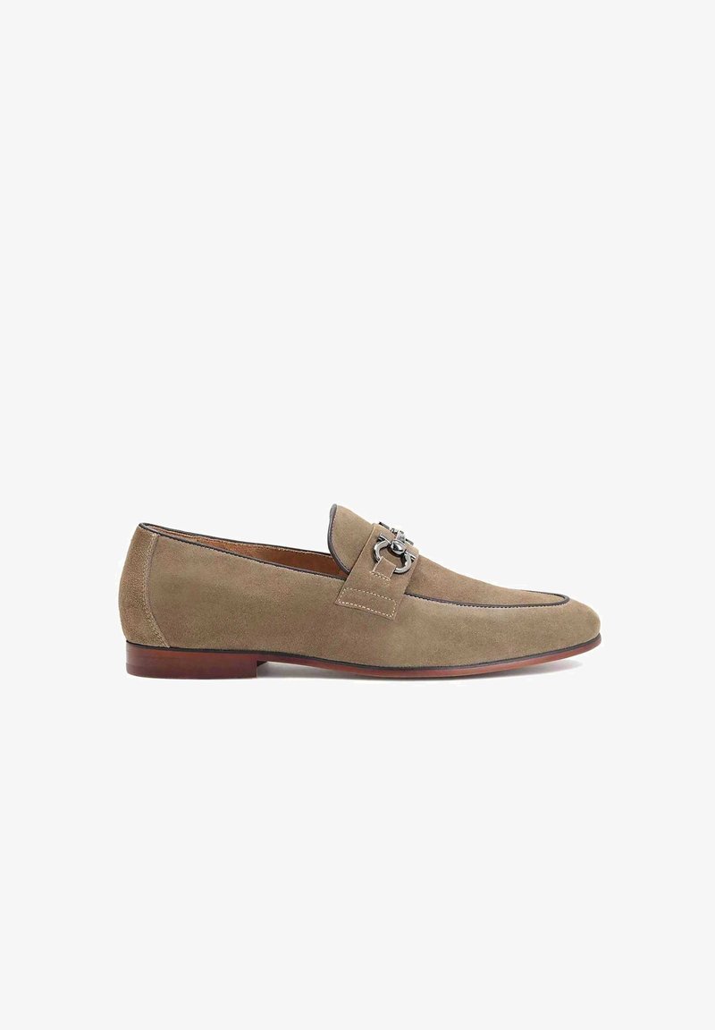 Mocassin beige en daim pour homme avec détail en métal argenté en forme de mors de cheval, semelle marron et bout pointu, présenté de profil sur fond blanc.